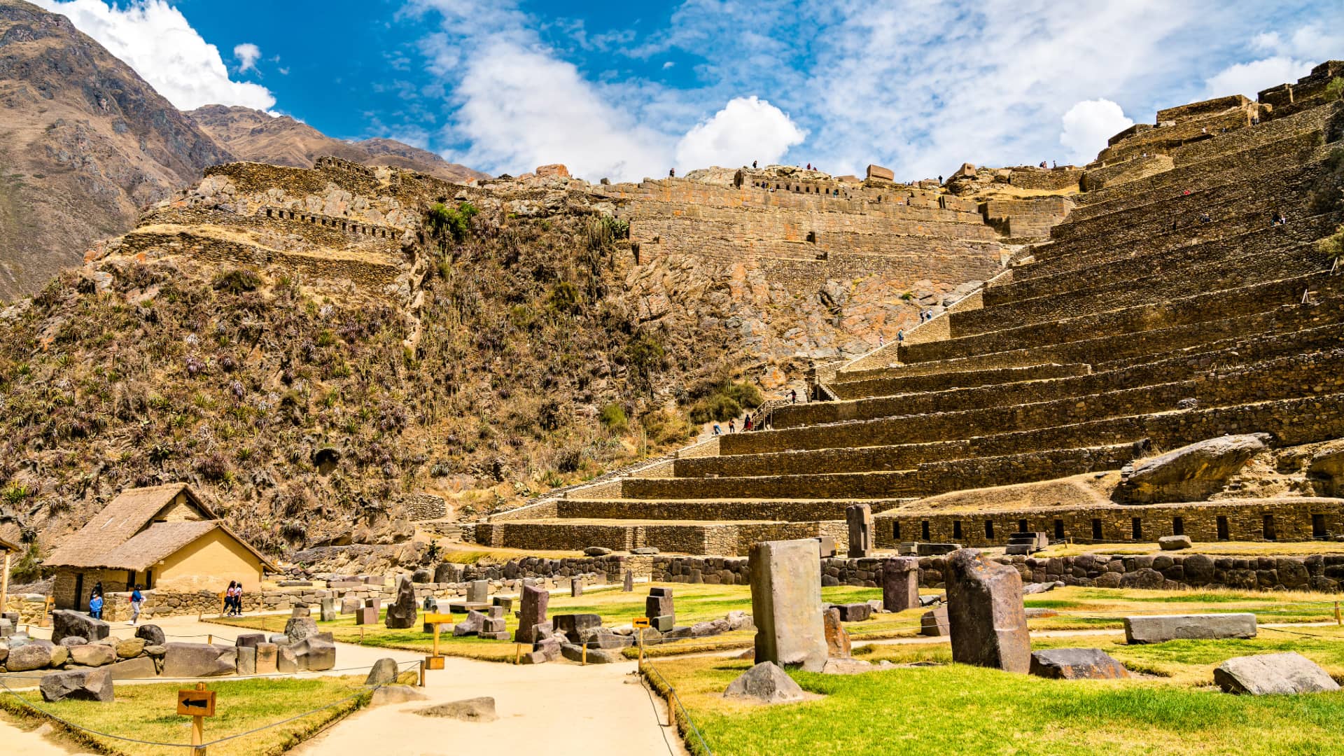 A wide shot of the grand Ruins of Ollantaytambo, featuring the large agricultural terraces, an ancient plaza with scattered stones, and the towering Inca fortress structure under a partly cloudy sky - Magical Cusco Travel Agency.