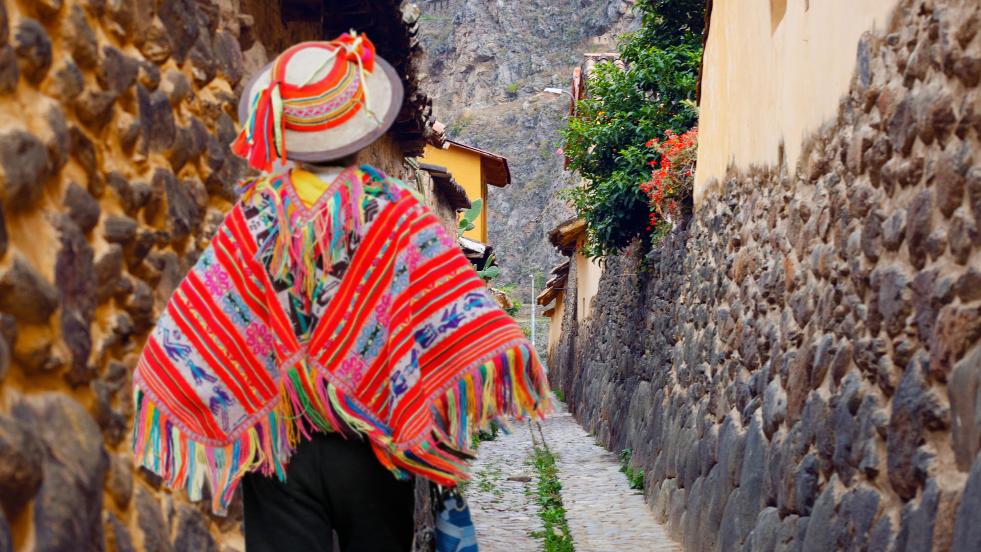 A person wearing a vibrant, traditional Andean poncho and hat walks through a narrow, ancient stone alleyway in the village near the Ruins of Ollantaytambo, with mountains in the background - Magical Cusco Travel Agency.