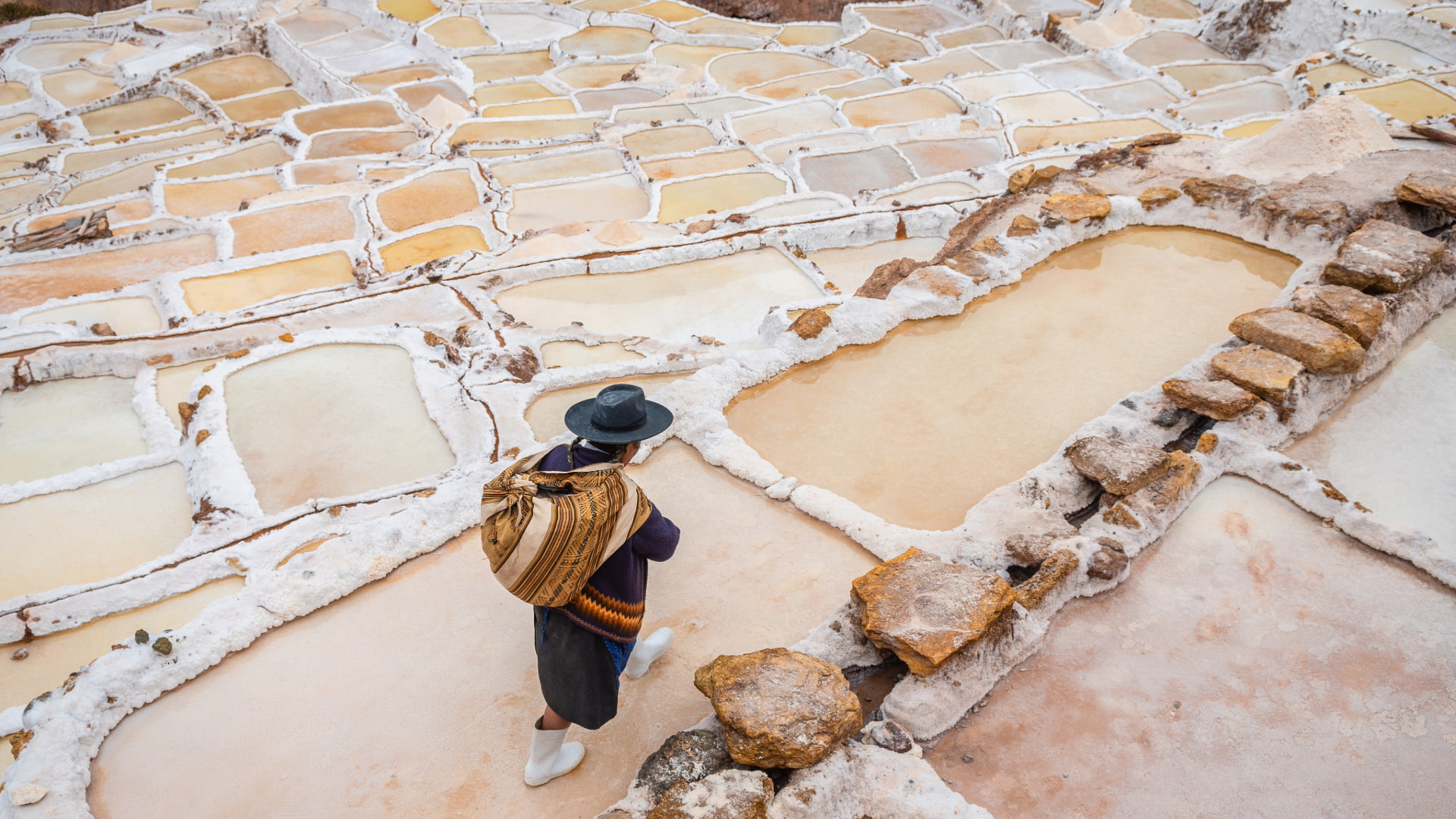 A tourist wearing a hat looks out over the vast, terraced salt ponds of Maras, with the dramatic Andean mountains in the background, a short distance from the Ruins of Ollantaytambo - Magical Cusco Travel Agency.