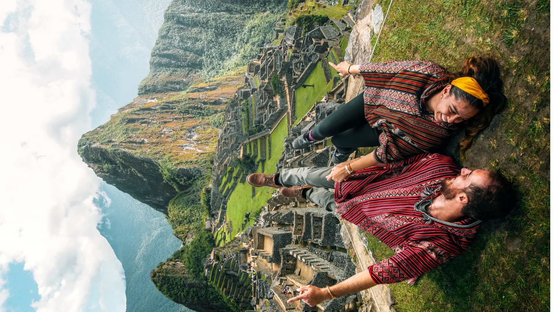 A happy couple lying on the grass and pointing towards the Machu Picchu citadel, capturing a fun moment while admiring the great Face of Machu Picchu. - Magical Cusco Travel Agency