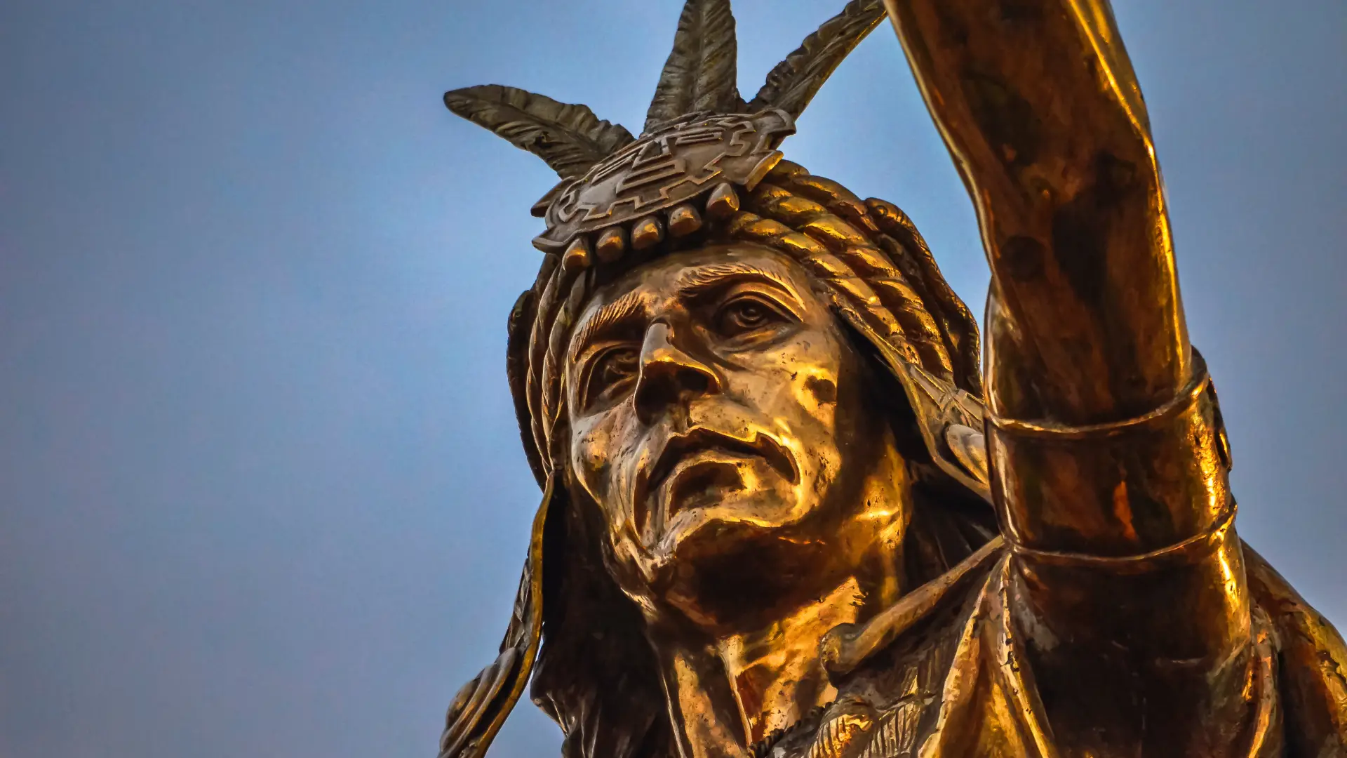 A close-up of a golden statue depicting an Inca figure wearing a feathered headdress, set against a blurred sky. - Magical Cusco Travel Agency