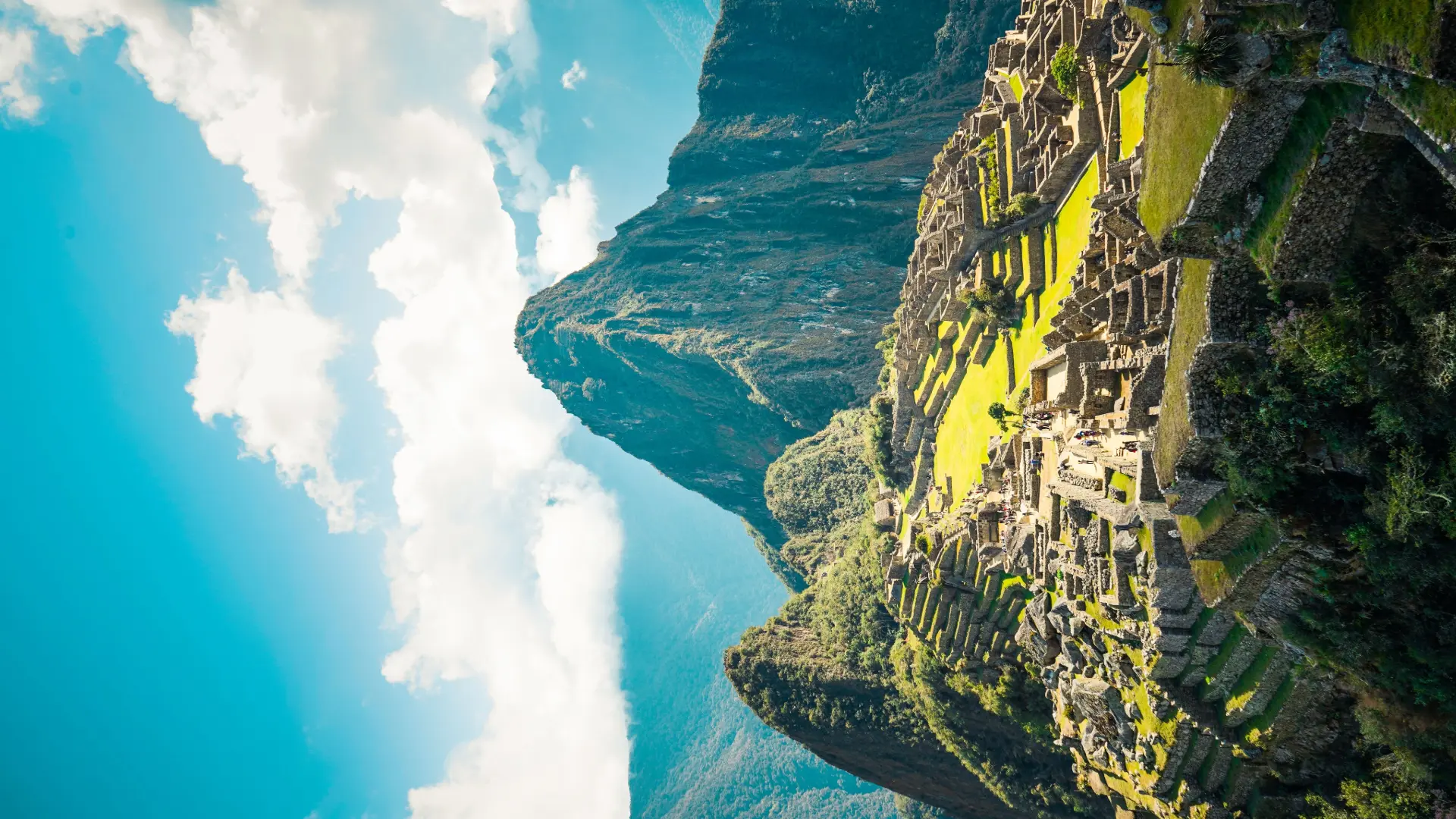 A side-angle panoramic view of the Machu Picchu citadel and Huayna Picchu peak under a bright blue sky, showing the iconic landscape of the Face of Machu Picchu. - Magical Cusco Travel Agency
