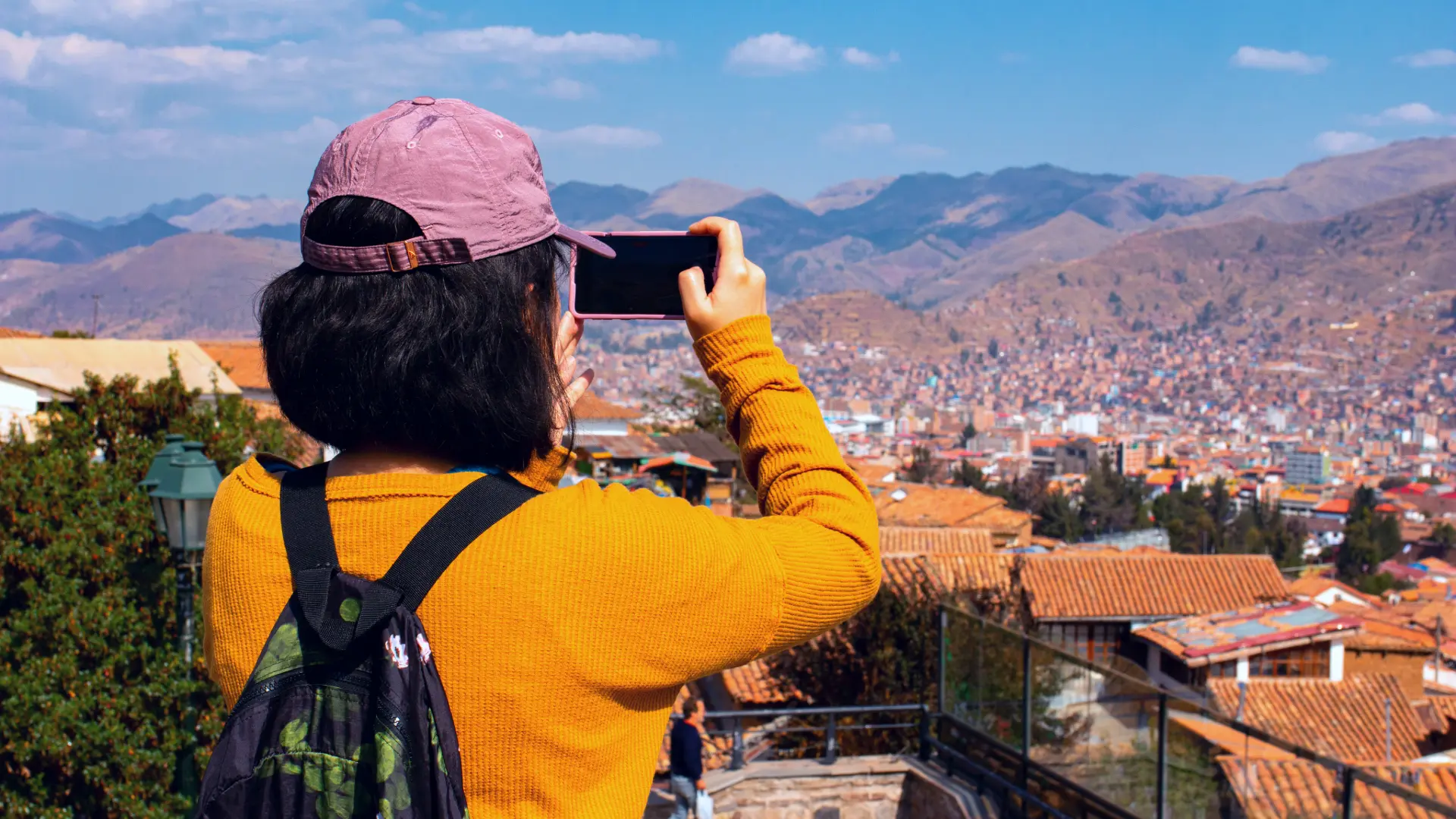 A tourist with a baseball cap and yellow sweater taking a photo of the Cusco cityscape with a smartphone, capturing a panoramic view of the red-roofed buildings and surrounding mountains. - Magical Cusco Travel Agency