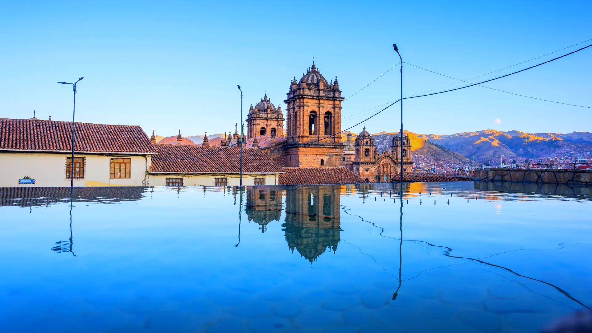 A serene view of a historical church in Cusco at sunset, beautifully reflected in a still pool of water with the city and mountains in the background. - Magical Cusco Travel Agency
