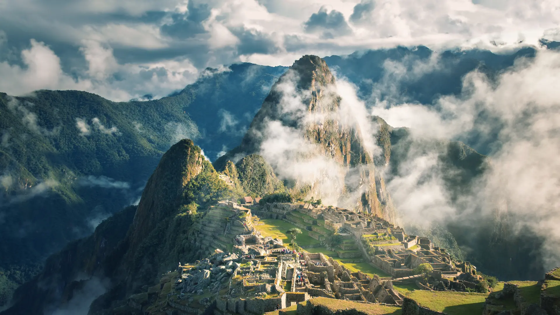 A breathtaking aerial view of the ancient Inca citadel of Machu Picchu, partially shrouded in clouds and mist, nestled amidst towering green mountains. - Magical Cusco Travel Agency