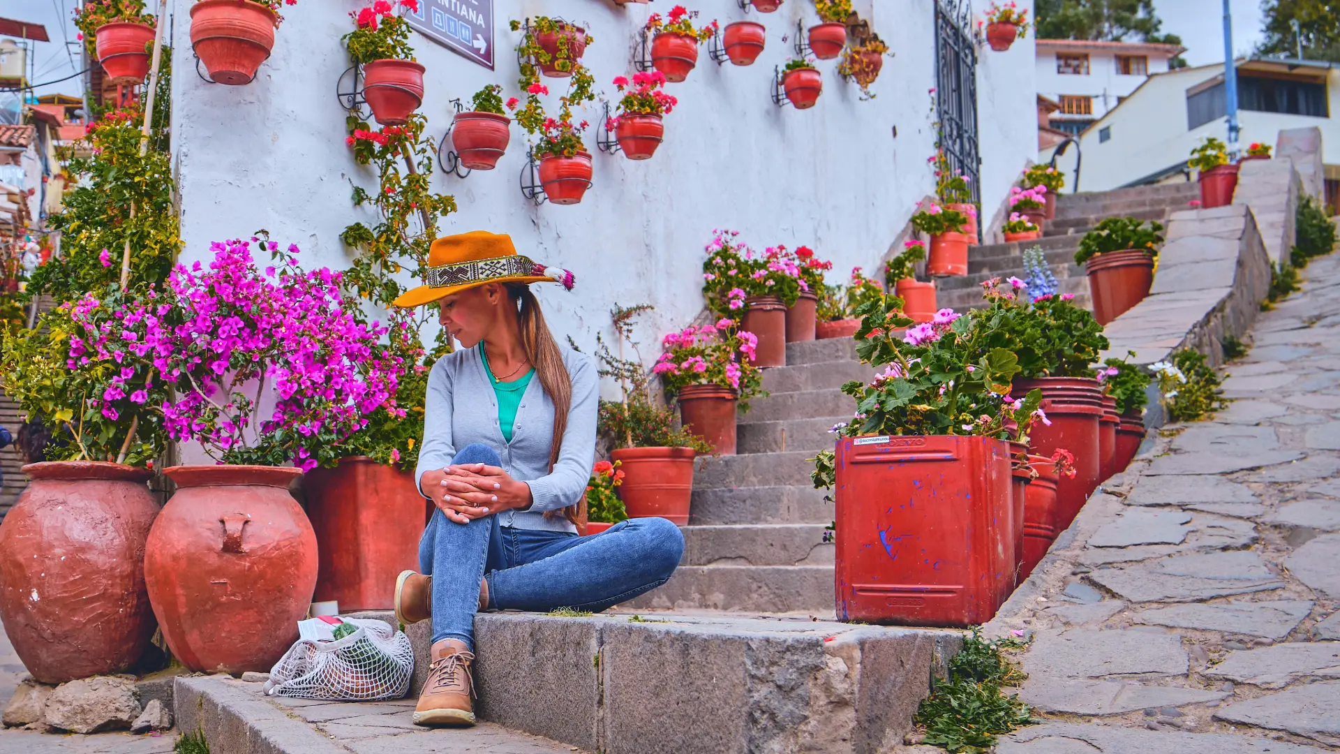 A female tourist in a hat and jeans sits on stone steps decorated with potted plants and flowers, enjoying a peaceful moment while Walking Cusco. - Magical Cusco Travel Agency