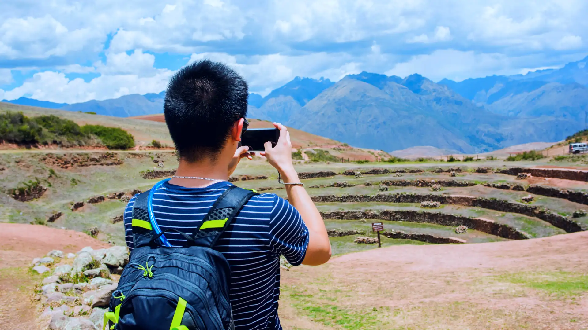 A tourist with a backpack is taking a photo of the ancient Inca agricultural terraces of Moray, part of the Moray and Maras Salt Mines tour. - Magical Cusco Travel Agency