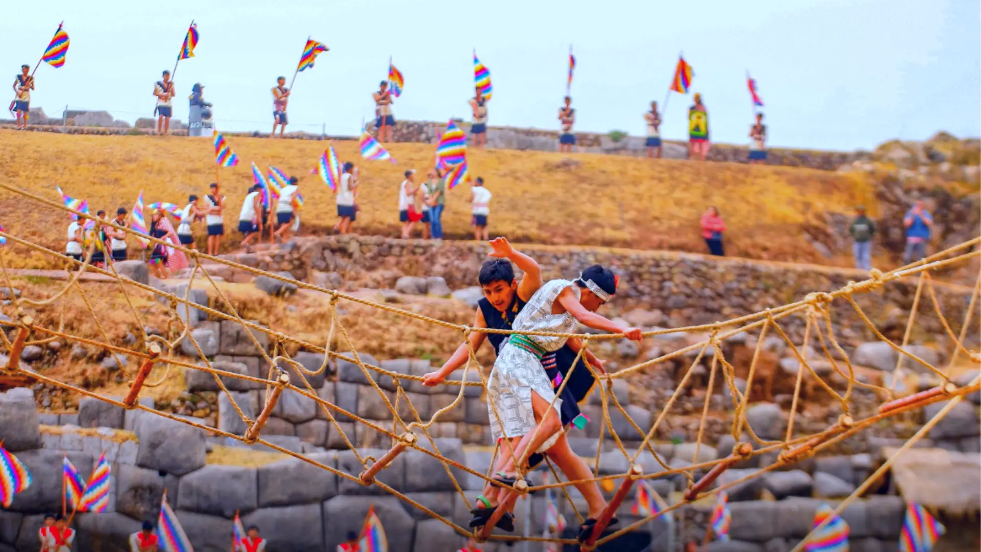 Young Peruvians participating in the Warachikuy Inca Ritual, crossing a rope bridge while spectators cheer them on with Andean flags. - Magical Cusco Travel Agency