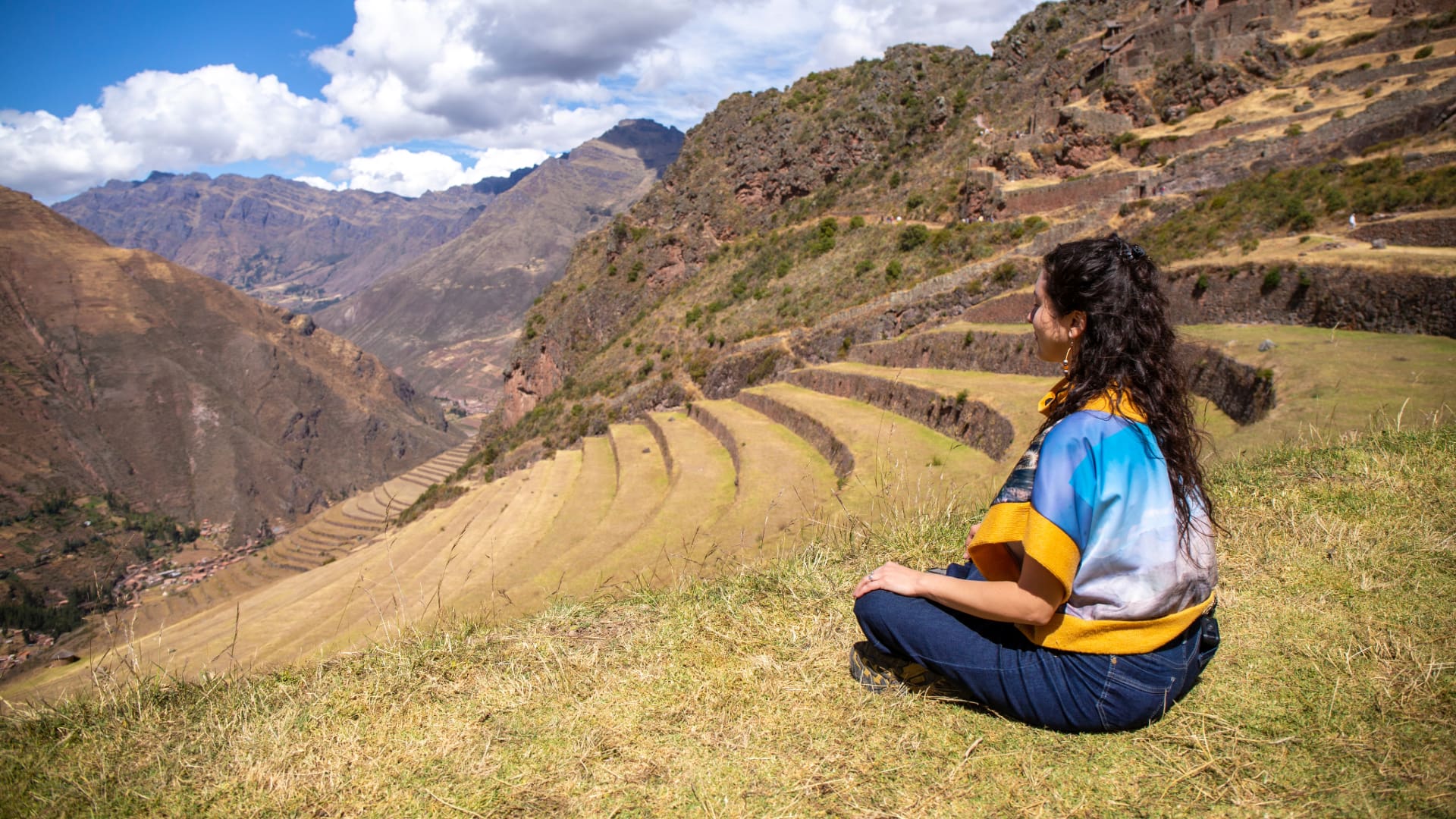A woman sits on a grassy slope, looking out over the layered, curved Inca Terraces and the deep Sacred Valley below. - Magical Cusco Travel Agency