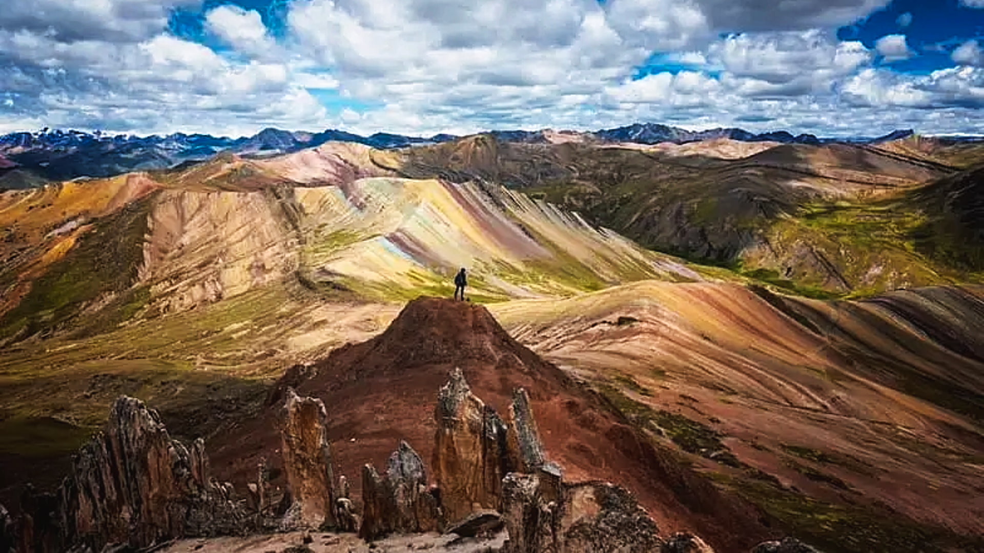 Person standing on a rocky outcrop overlooking the multicolored terrain of Palccoyo Rainbow Mountain – Magical Cusco Travel Agency