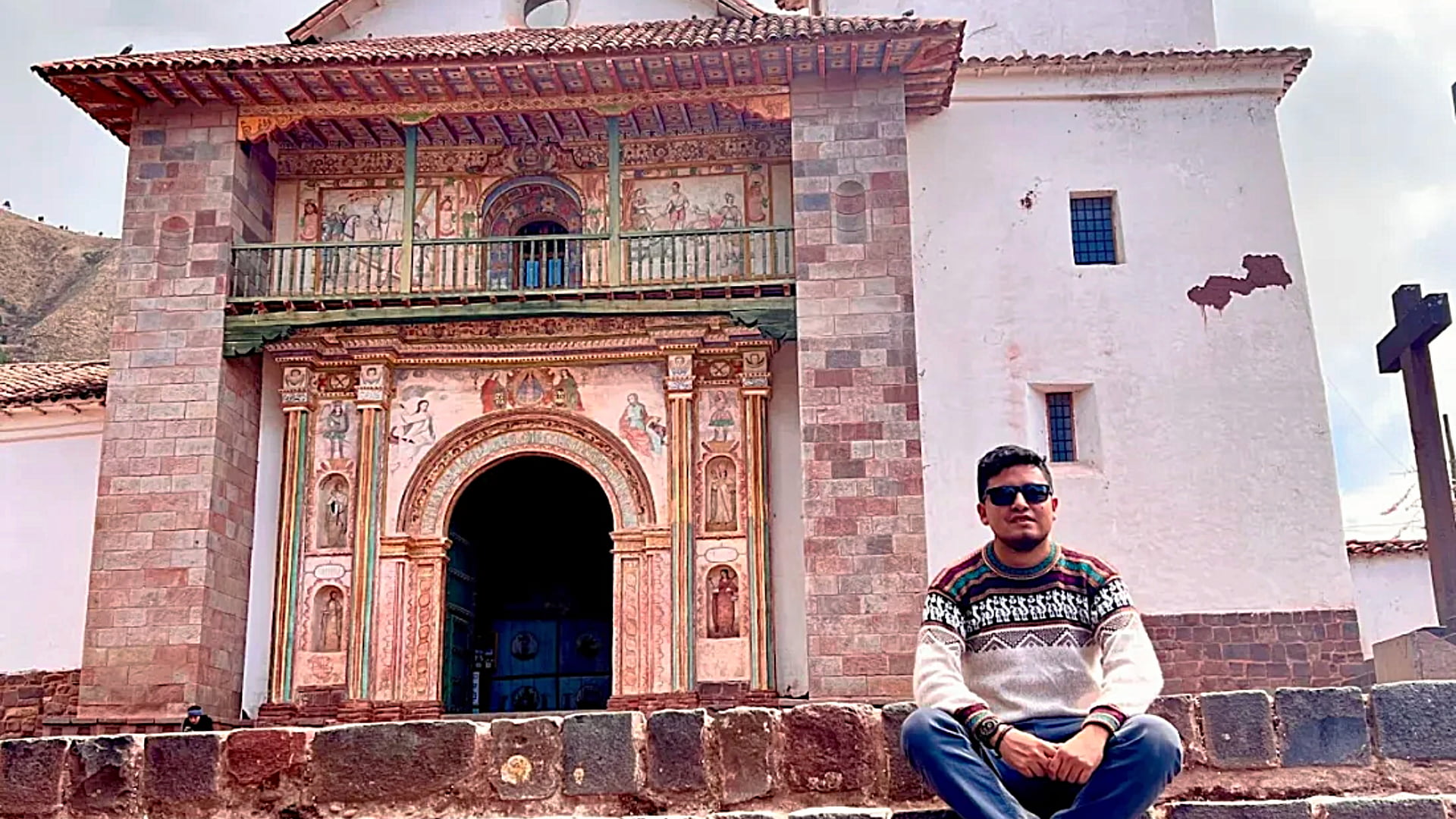A male traveler sitting on the steps in front of the highly decorated, Baroque facade of the Andahuaylillas Temple, a site central to local Andahuaylillas traditions and culture. - Magical Cusco Travel Agency