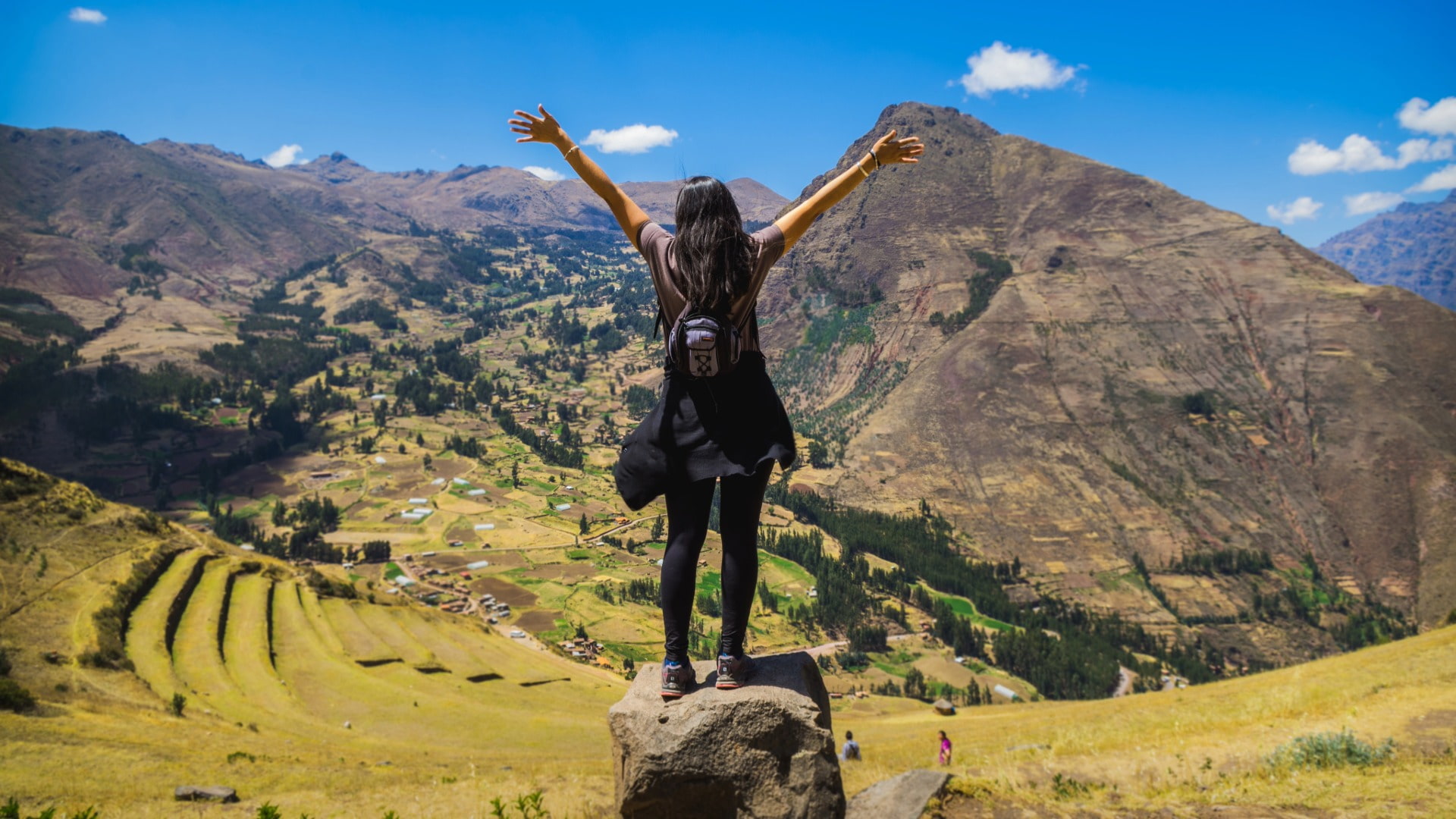A woman stands with her arms raised on a rock, enjoying a panoramic view of the agricultural terraces and majestic mountains of the Sacred Valley. - Magical Cusco Travel Agency